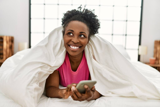 African american woman using smartphone lying on bed at bedroom - Photo, image