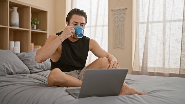 Young hispanic man using laptop drinking coffee at bedroom - Photo, Image