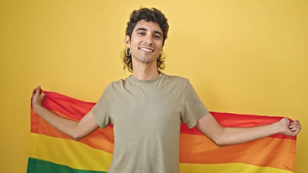 Giovane uomo ispanico sorridente fiducioso tenendo bandiera arcobaleno su isolato sfondo giallo - Foto, immagini