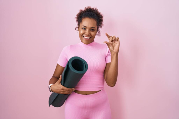 Junge hispanische Frau mit lockigem Haar hält Yogamatte über rosa Hintergrund lächelnd und selbstbewusst gestikulierend mit der Hand und macht ein kleines Zeichen mit den Fingern und der Kamera. Maßnahmenkonzept.  - Foto, Bild