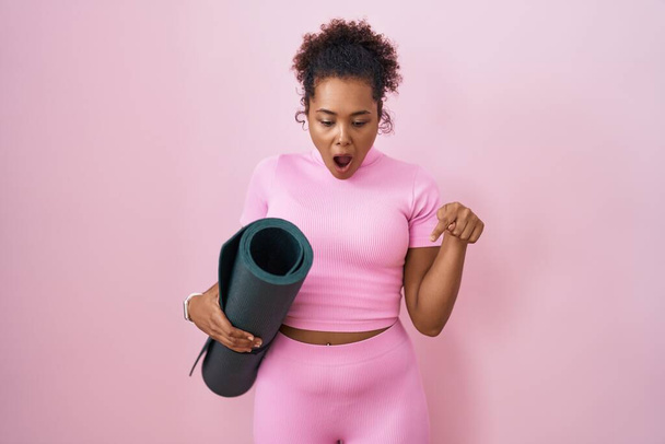 Junge hispanische Frau mit lockigem Haar hält Yogamatte über rosa Hintergrund und zeigt mit Fingern nach unten, die Werbung zeigen, überraschtes Gesicht und offener Mund  - Foto, Bild