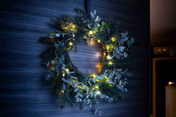 Corona de Navidad de pino y eucalipto forrajeado, con luces de cuerda iluminadas, colgando de la puerta de apertura. - Foto, imagen