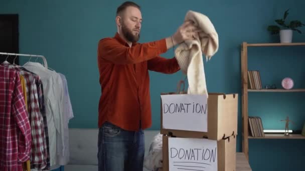Όμορφος άντρας συσκευάζει κουτί με ρούχα στο σαλόνι του στο σπίτι. Έννοια δωρεάς ρούχων - Πλάνα, βίντεο
