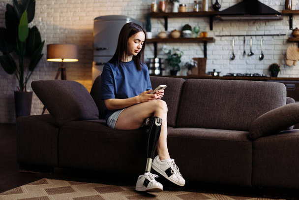 Женщина с ограниченными возможностями сидит на диване и печатает сообщения на смартфоне. Привлекательная леди с протезной ногой активно болтает с парнем по мобильному телефону - Фото, изображение