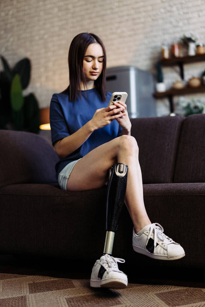 Kobieta niepełnosprawna siedzi na kanapie i pisze wiadomości na smartfonie. Atrakcyjna kobieta z protezą nogi zaangażowana w ożywioną rozmowę z chłopakiem przez telefon komórkowy - Zdjęcie, obraz