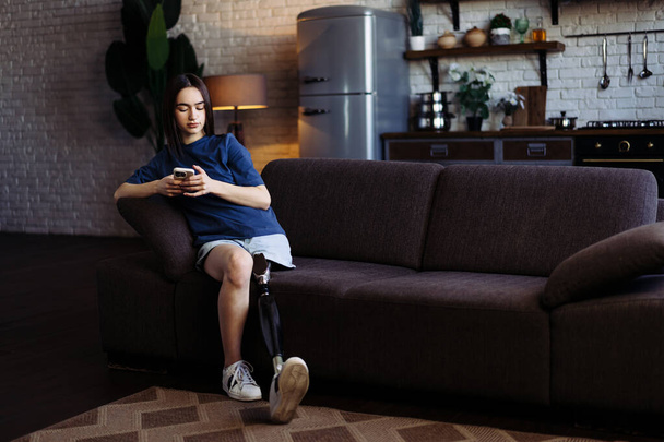 Nuori nainen, jolla on vamma, kertoo tarinoita ystävälleen puhelimitse. Lady proteesin jalka rento pukea istuu pehmeällä sohvalla ja tyypit viestejä älypuhelimeen - Valokuva, kuva