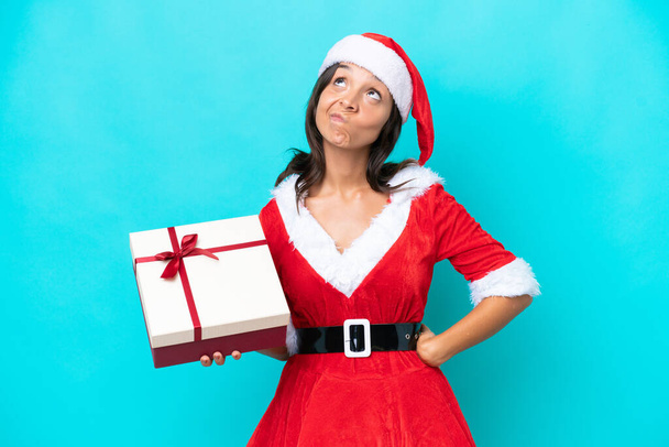 Anne Noel gibi giyinmiş genç İspanyol kadın elinde mavi arka planda izole edilmiş bir hediye tutuyor ve yukarı bakıyor. - Fotoğraf, Görsel