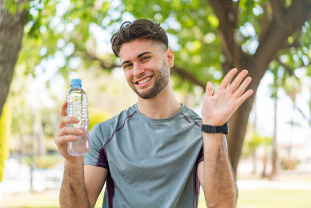 młody przystojny sport człowiek z butelką wody na świeżym powietrzu salutując z ręki z szczęśliwy wyraz twarzy - Zdjęcie, obraz