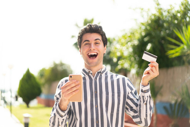 Молодой красивый мужчина покупает с мобильного и держит кредитную карту с удивленным выражением лица - Фото, изображение