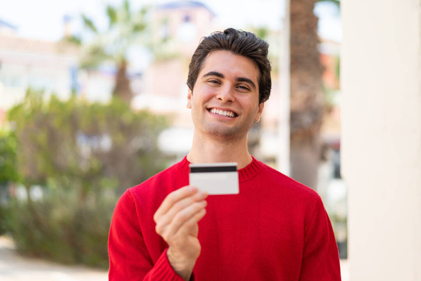 Junger gutaussehender Mann hält im Freien mit glücklichem Gesichtsausdruck eine Kreditkarte in der Hand - Foto, Bild