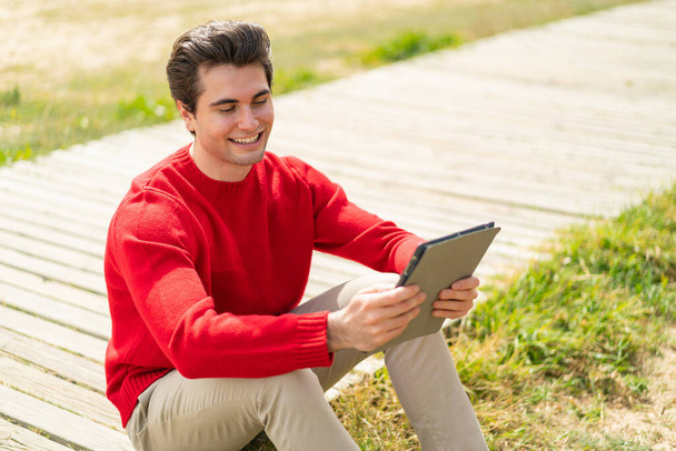 Jonge knappe man met een tablet in de buitenlucht met vrolijke uitdrukking - Foto, afbeelding