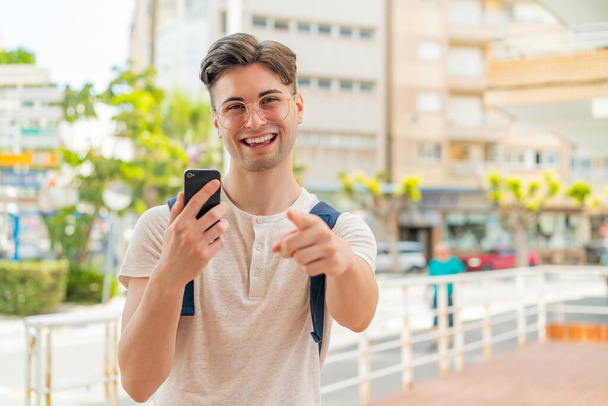 Молодой красивый мужчина, используя мобильный телефон на открытом воздухе указывает пальцем на вас с уверенным выражением - Фото, изображение