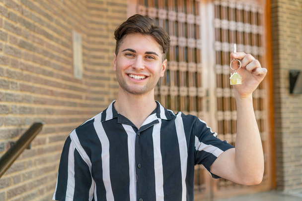 Νεαρός όμορφος άντρας που κρατάει τα κλειδιά του σπιτιού στην ύπαιθρο χαμογελώντας πολύ - Φωτογραφία, εικόνα