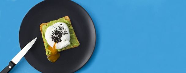 Sandwich mit pochiertem Ei und Avocado auf dem schwarzen Teller auf blauem Hintergrund. Ansicht von oben. Kopierraum. - Foto, Bild
