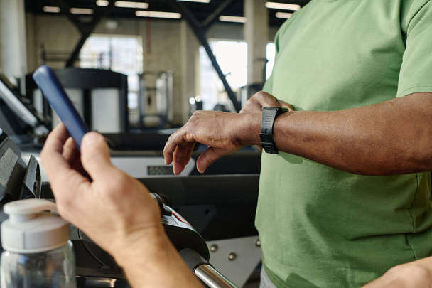 Hombre negro desconocido mirando su reloj con otra persona caucásica sosteniendo el teléfono inteligente en la mano - Foto, imagen