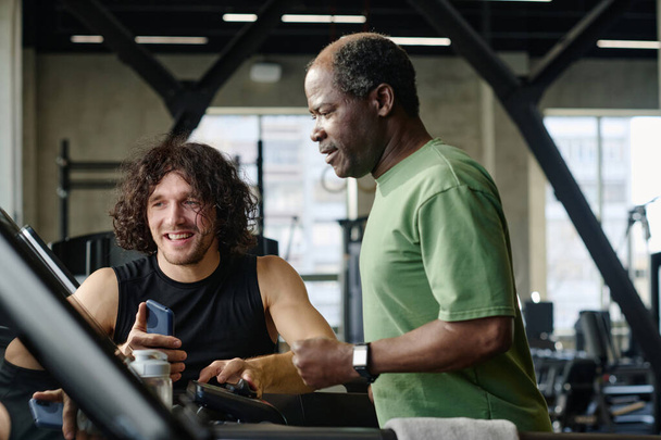 Yaşlı Afro-Amerikan adam ve Kafkas atleti spor salonundaki koşu bandında bilgi kontrol ediyorlar. - Fotoğraf, Görsel