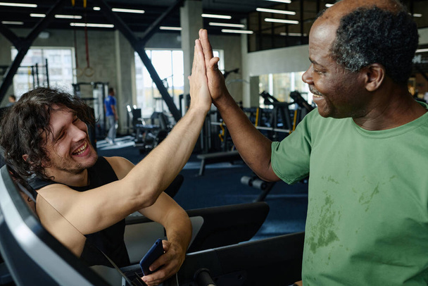 Улыбающийся пожилой черный мужчина и счастливый белый парень, дающий друг другу пять в фитнес-центре - Фото, изображение