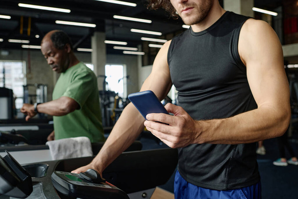Unknown caucasian guy walking on treadmill with smartphone, black gym goer working out on background - Foto, afbeelding