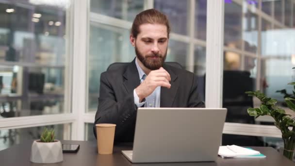 Homme barbu pensif utilisant la technologie sans fil pour le travail en ligne dans le bureau. Expert caucasien concentré avec ordinateur portable et tasse de café sur son bureau compte tenu des tâches et dactylographier sur le clavier. - Séquence, vidéo