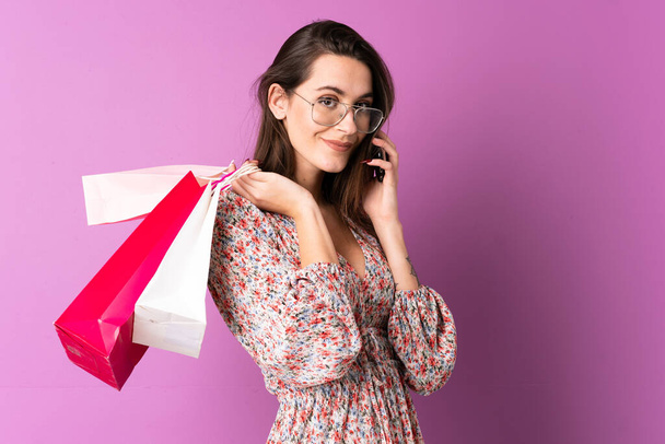 Giovane donna su sfondo viola isolato tenendo borse della spesa e chiamando un amico con il suo telefono cellulare - Foto, immagini