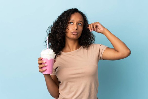 tiener meisje met aardbei milkshake geïsoleerd op blauwe achtergrond met twijfels en denken - Foto, afbeelding