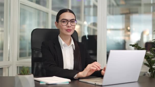 Concentrated entrepreneur wearing stylish classic clothes sitting in office and working with modern laptop. Beautiful successful female with dark hair smiling and putting hands on table indoor. - Footage, Video