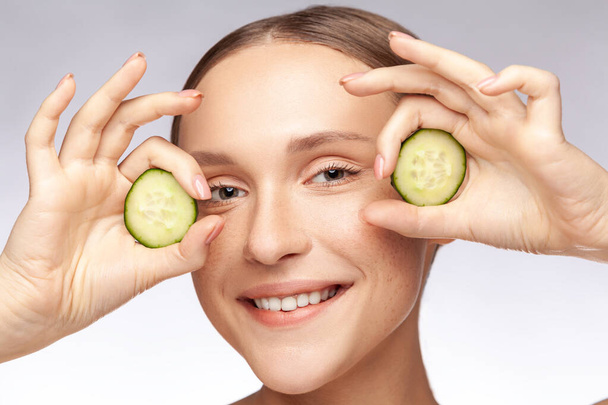 Closeup portrait of adorable beautiful woman taking care of her skin making cucumbers mask smiling to camera. Indoor studio shot isolated over gray background. - Foto, imagen