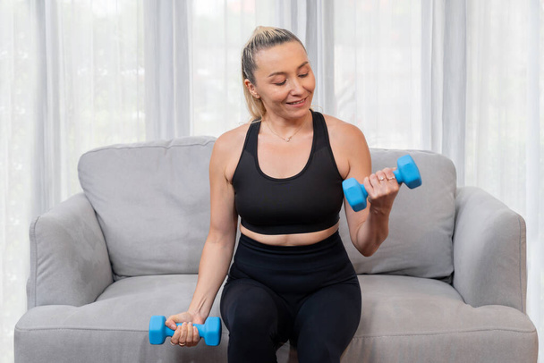 Sportos és sportos idős nő vesz részt a testedzés rutin, ül a kanapén, és emelés súlyzó otthon, mint koncepció az egészséges fit test testsúly életmód nyugdíjazás után. Kömény - Fotó, kép