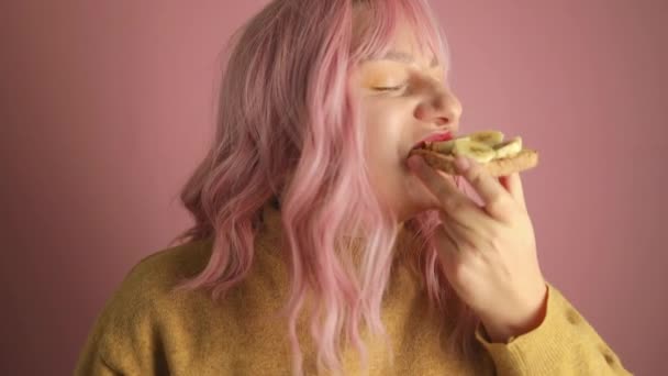 Stúdió portré boldog hangry rózsaszín haj nő enni ízletes szendvics banán és nézi a kamera álló rózsaszín elszigetelt háttér.  - Felvétel, videó