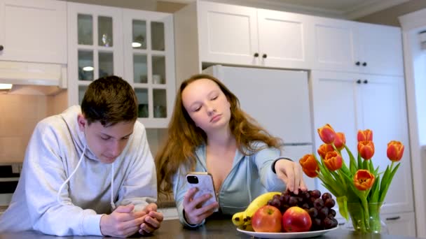 adolescentes en la cocina comiendo frutas jugando en el teléfono hermosos muebles blancos Acogedor hogar. plato con manzanas y plátanos Flores en la mesa de vacaciones un día ordinario en América - Imágenes, Vídeo