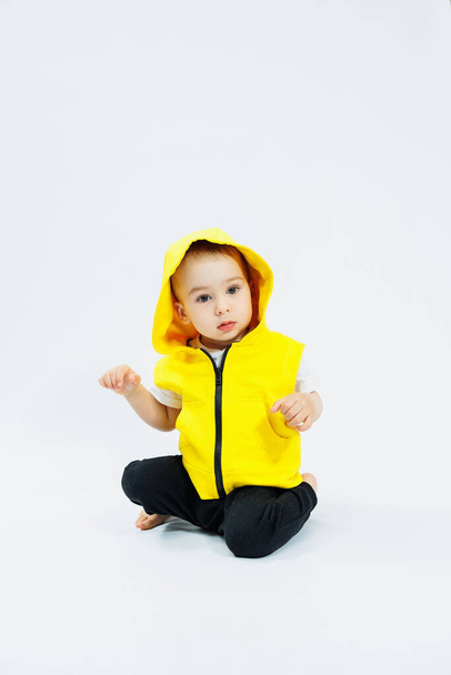 Kleine vrolijke jongen van 1 jaar oud in een geel vest op een witte achtergrond. Heldere kleren voor kinderen. Portret van een vrolijk kind in een kap - Foto, afbeelding