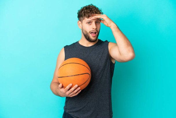 Bonito jovem jogando basquete isolado no fundo azul fazendo gesto surpresa enquanto olha para o lado - Foto, Imagem