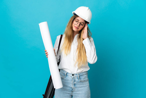 Giovane architetto donna con casco e tenendo cianografie isolate su sfondo blu con mal di testa - Foto, immagini