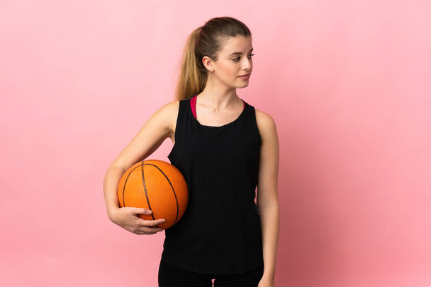 Mujer rubia joven jugando baloncesto aislado sobre fondo rosa mirando hacia un lado - Foto, Imagen