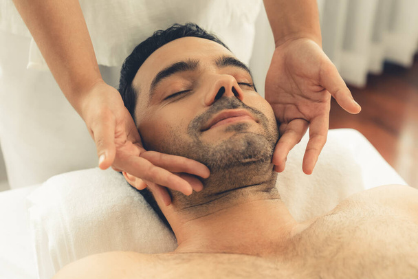 Кавказский мужчина наслаждается расслабляющим анти-стрессовым массажем головы и расслабляющим отдыхом кожи лица в дневном спа современном светлом окружении на роскошном курорте или в спа-салоне отеля. Quiescent - Фото, изображение