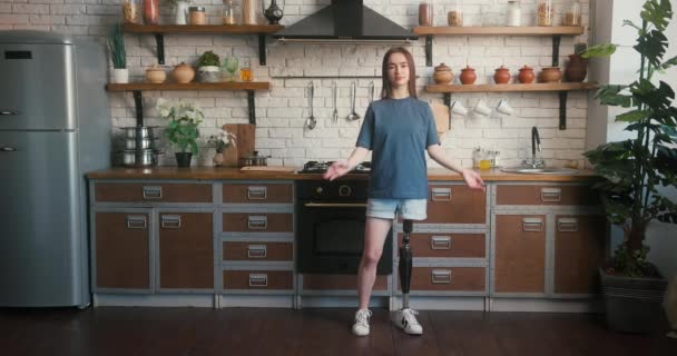 Μελαχρινή γυναίκα με τεχνητό άκρο στέκεται στην κουζίνα με σταυρωμένα χέρια. Γυναίκα με αναπηρία αποπνέει αυτοπεποίθηση ποζάροντας για την κάμερα στην άνετη κουζίνα - Πλάνα, βίντεο