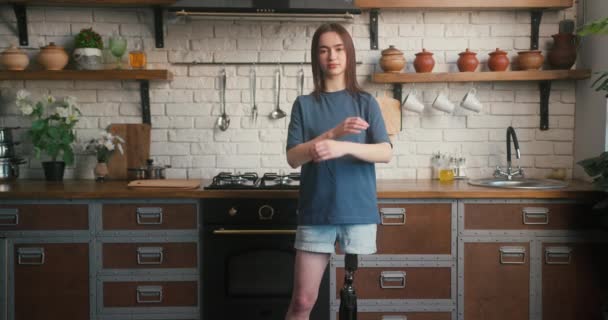 Brünette Frau mit künstlichen Gliedmaßen steht mit verschränkten Armen in der Küche. Behinderte Frau strahlt Zuversicht aus, posiert in gemütlicher Küche für die Kamera - Filmmaterial, Video
