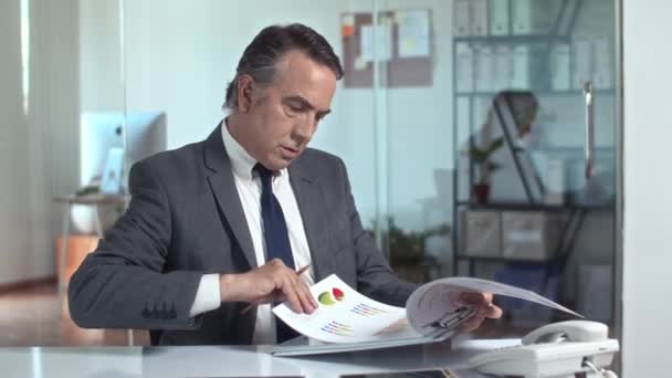 Keskikokoinen laukaus kypsä miesyrittäjä tekee muokkauksia raportissa istuu pöydällä modernissa toimistossa - Materiaali, video