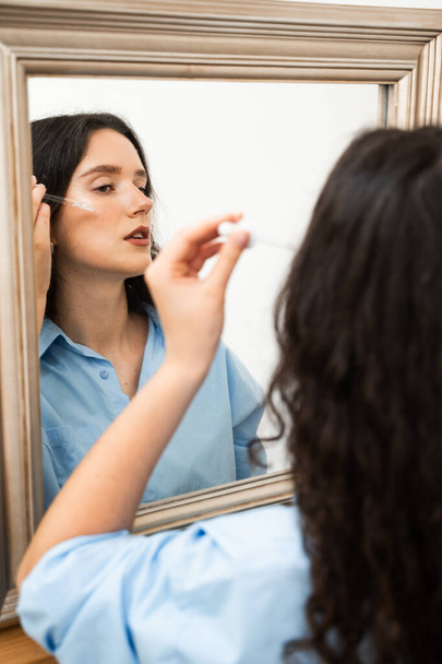 Κορίτσι με υαλουρονικό οξύ ή πιπέττα ορού στα χέρια κοιτάζει στον καθρέφτη. Νεαρή γυναίκα εφαρμόζει ενυδατικό ορό στο πρόσωπο του δέρματος - Φωτογραφία, εικόνα