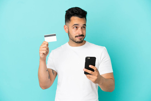 Giovane uomo caucasico isolato su sfondo blu che acquista con il cellulare con una carta di credito mentre pensa - Foto, immagini