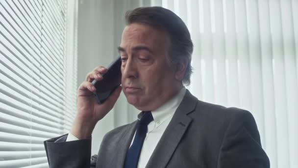 Střední záběr senior finančního manažera volat klienta pomocí smartphonu, zatímco stojí vedle okna v kanceláři - Záběry, video