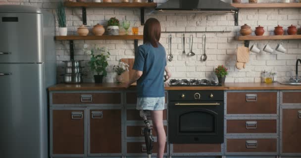 Женщина с протезной ногой ставит горшок на газовую плиту для приготовления пищи. Молодая женщина с ограниченными возможностями эксперименты с новыми рецептами в современной кухне на дому - Кадры, видео