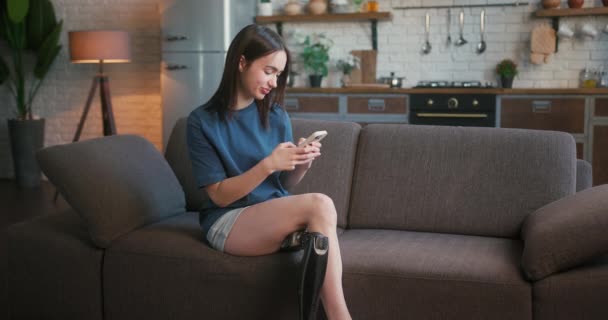 Woman with disability sits on sofa and types messages on smartphone. Attractive lady with prosthetic leg engaged in lively chat with boyfriend on mobile phone - Footage, Video