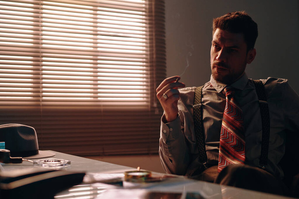 Jonge penetrante zakenman in retro formalwear die een sigaret vasthoudt terwijl hij aan tafel zit in donker kantoor en aan iets denkt - Foto, afbeelding