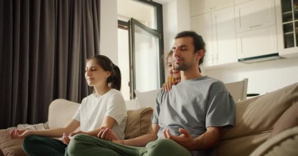 A brunette girl in a white T-shirt together with her husband, a brunette man in a gray T-shirt, meditates while sitting on light brown daughter girl, a little brunette girl in a yellow dress - Záběry, video