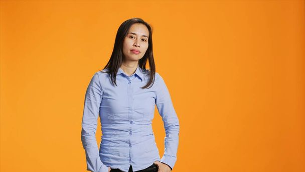 Portret van zorgeloos Aziatisch meisje poserend met zelfvertrouwen, met een blauw shirt aan en vol zelfvertrouwen in de studio. Mooie filipino persoon staande over oranje achtergrond, glimlachend met elegantie. - Foto, afbeelding