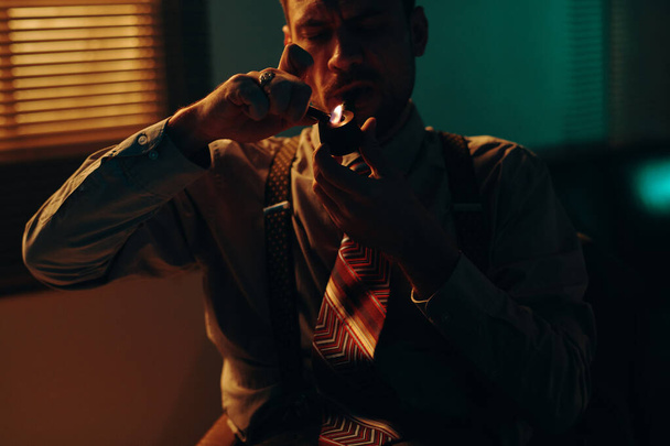 Jeune homme d'affaires ou policier en tenue de cérémonie allumant la pipe à tabac assis dans un bureau sombre devant la caméra et fumant - Photo, image