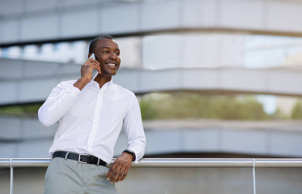 Portrét Usmívající se černý podnikatel mluví na mobilním telefonu, zatímco stojí venku v blízkosti moderního kancelářského centra, Happy African American Male Entrepreneur těší mobilní telefon konverzace, kopírovat prostor - Fotografie, Obrázek
