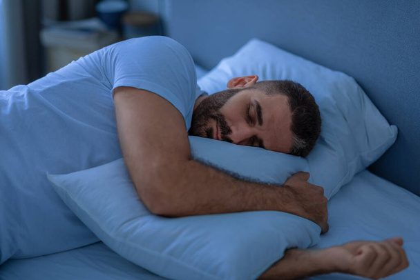 Rauhallinen kaveri nukkuu syleilevä tyyny makaa sängyssä kotona yöllä. Lepää rutiini, torkut ja vapaa-aika. Vuosituhannen mies lepää rauhassa kodikas moderni makuuhuone. Heikko valo - Valokuva, kuva
