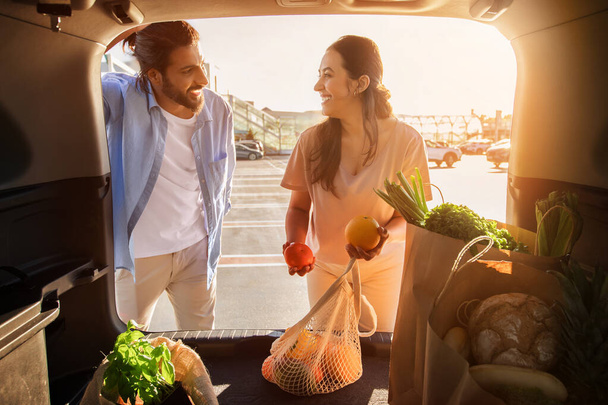 Νεαρό ζευγάρι της Μέσης Ανατολής μοιράζεται τη στιγμή της χαράς, ενώ επιθεώρηση φρέσκα πιπεριές και πορτοκάλια από το πορτ-μπαγκάζ του αυτοκινήτου τους στο πάρκινγκ - Φωτογραφία, εικόνα
